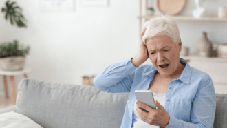 older woman in a blue shirt holding her head with one hand and a smartphone in the other